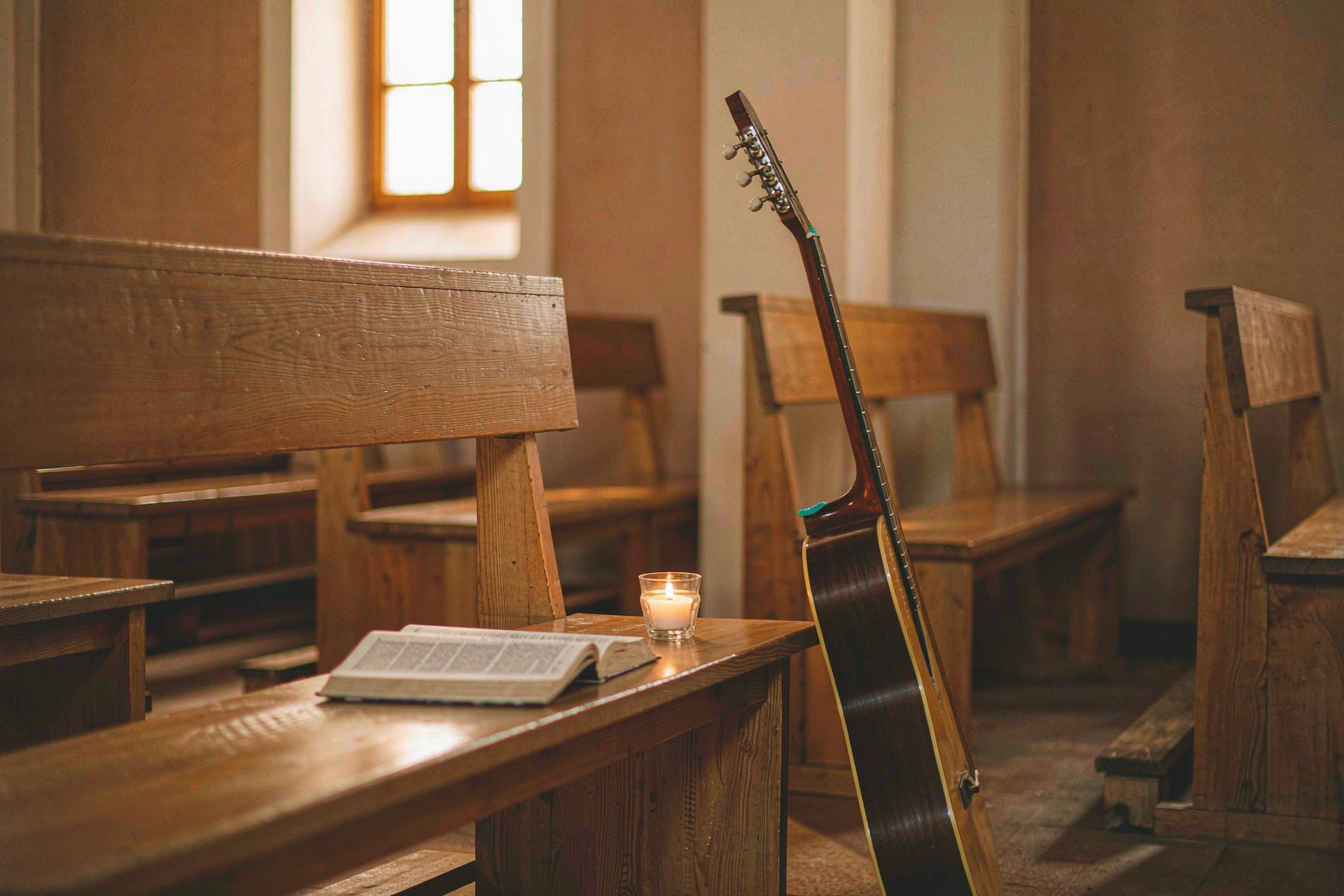 An open hymnal on a chapel pew beside an acoustic guitar and a single candle in golden light