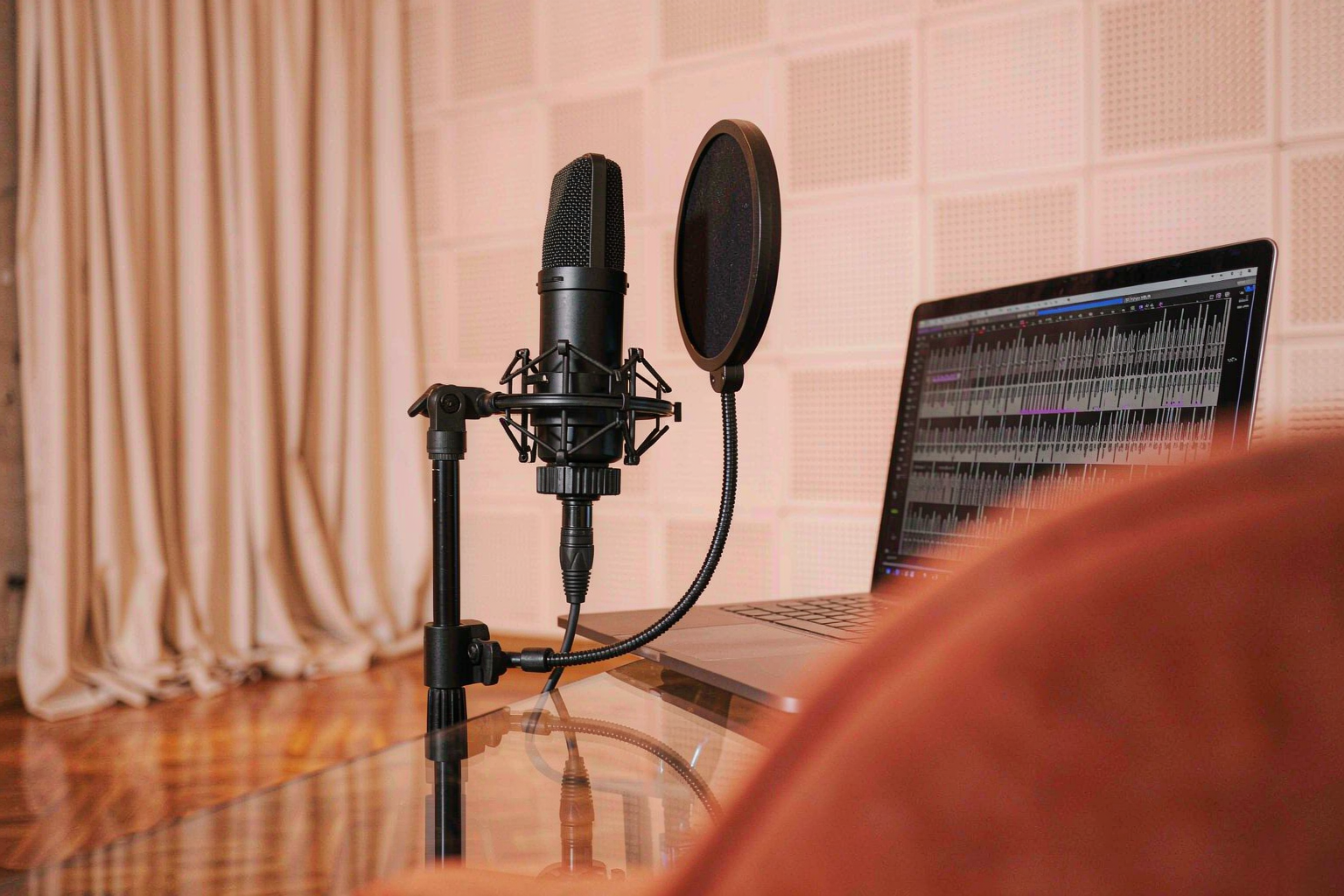 A condenser microphone and pop filter in a bright vocal booth with a laptop showing a waveform